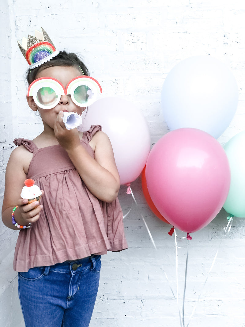 A little girl wearing a birthday crown and rainbow glasses standing next to helium filled balloons 